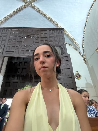 A medium shot shows a woman in a yellow dress indoors. In the background is an ornately carved wooden door and ceiling decorations. The location is likely a church in Orizaba, Mexico. Two other women can be seen behind the main subject, and a man in a suit is partially visible at the bottom.