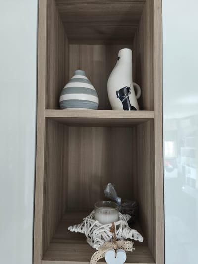 The image shows a close-up view of a light wood-grained shelf unit with two open compartments. The upper compartment contains two decorative vases. On the left is a cylindrical vase with horizontal grey and white stripes. To its right is a tall, off-white vase with abstract black markings and a handle. The lower compartment contains a glass jar with a white candle inside, resting on a white woven star-shaped object. A small, white heart-shaped ornament with lace ribbon is attached to the woven star. The background appears to be a white cabinet or wall. The scene is indoors. The provided information about Passignano sul Trasimeno, Italy is not relevant to the visual content of the image.