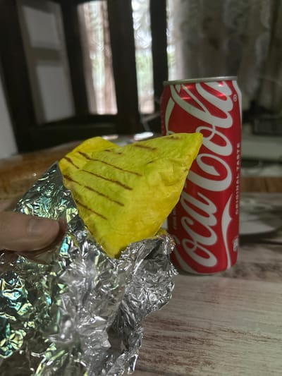 An overhead, close-up shot shows a hand holding a partially unwrapped, grilled yellow wrap. The wrap has diagonal grill marks and is mostly obscured by crinkled aluminum foil. To the right of the wrap is a red Coca-Cola can with its distinctive white logo. The objects are placed on a wooden table. In the blurred background, a window with wooden mullions reveals a soft, natural light. The scene appears to be indoors.