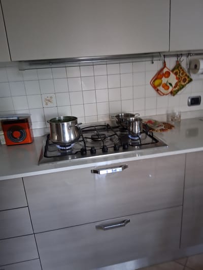 This media file shows a close-up view of a kitchen scene. The primary subjects are kitchen objects, including a stovetop with a pot on it, and a wall with tiled backsplashes and decorative items. The stovetop is made of stainless steel and has four burners, one of which is lit, with a blue flame visible under a silver pot. The pot has a rounded shape and handles. Above the stovetop, hanging from a rack, are two decorative potholders. One is patterned with sunflowers, and the other features images of oranges and a lemon. On the wall behind these items are white square tiles, some with a faint floral pattern. To the right of the stovetop are kitchen cabinets with modern, silver handles. An orange kitchen timer is visible on a shelf below the stovetop. The lighting suggests it is daytime, possibly in a home kitchen. There are no people visible in the image. The location context is Padua, Italy. No visible text is present in the image.