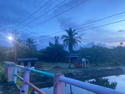 The image depicts a serene twilight scene in Aneuk Paya, Indonesia. A bridge railing, painted in vibrant pastel colors of pink, purple, and teal, dominates the foreground. Beyond the bridge, a tranquil body of water reflects the dusky sky. The midground features a small, dark-colored building with a metal roof and two white doors, flanked by lush green vegetation. Further back, several palm trees and other tropical foliage create a dense backdrop, interspersed with a few structures, including a red gazebo with a dark roof and an orange-hued building with a dark roof. Power lines stretch across the sky, one pole illuminated by a bright light. The sky itself is a canvas of deep blues and grays, with scattered clouds hinting at the fading daylight. The overall atmosphere is peaceful and rural, characteristic of a quiet evening in this Indonesian locality.