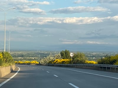 The image captures a view of a highway leading into a valley with a distant city skyline. The highway is paved, with white lane markings and a concrete barrier along the right side. There are several streetlights visible on the left, suggesting an urban or suburban setting. Along the roadside, yellow flowering bushes and green trees are abundant, adding color to the scene. In the distance, a vast expanse of land is covered with greenery, dotted with buildings and structures, indicating a populated area. A speed limit sign showing "110" is posted on the right side of the highway. The sky is partly cloudy with a mix of blue and white, suggesting a pleasant day. This scene is located in the city of Urbnisi, Georgia.