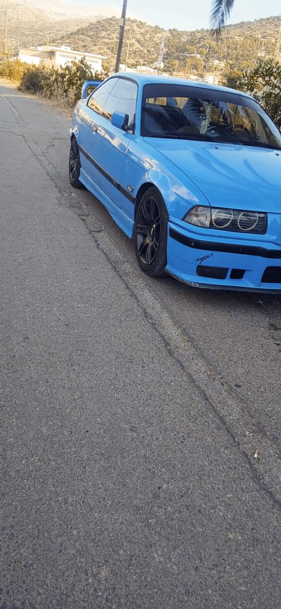 The image shows a bright blue BMW coupe parked on the side of a paved road. The car is angled with its front driver's side closest to the camera. It has black racing stripes along the side and black rims. The car is clean and appears to be in good condition.
The background suggests a Mediterranean setting with hills covered in vegetation and scattered buildings. The sun is shining, indicating it is daytime, and the weather appears to be clear. There is some greenery on the left side of the image, possibly trees or bushes. The road surface is asphalt with a visible crack running diagonally across it.
A portion of the car's windshield shows a reflection of palm fronds, hinting at the presence of palm trees nearby. On the front fender, there is a sticker that appears to say "AWAJ" in stylized lettering. No people are visible in the image. The overall impression is a still shot of a car parked in a scenic, possibly vacation-like, location.