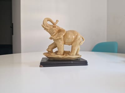 A light-colored, ivory-like figurine of an elephant stands on a dark rectangular base. The elephant is positioned as if mid-stride, with its trunk raised and curled upwards. It is placed on a white, glossy surface that appears to be a table. In the background, a white wall is visible, with a darker vertical element on the left side, possibly a doorway or a shadow. A bright teal-colored chair is partially visible to the right of the elephant, also in the background. The lighting suggests an indoor setting with diffused natural light. There is no indication of the specific location within the city of Katowice, Poland.