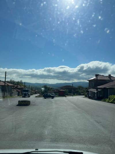 The image shows a road in Rish, Bulgaria. The road is paved and runs through a rural area with houses on either side. A tractor tire and a van are visible on the road. There are clouds in the sky and mountains in the distance. The sun is shining brightly.