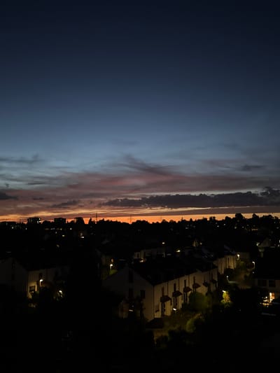 This is a nighttime aerial photograph of a residential area in Sindelfingen, Germany. The sky displays a gradient of deep blue to a dusky orange and pink, indicating twilight or a sunset afterglow. Below the sky, the silhouette of a densely packed neighborhood of houses and buildings is visible against the fading light. Many of the houses have their interior lights on, casting warm glows through the windows and illuminating some outdoor areas with streetlights. Trees and other greenery are present throughout the neighborhood, appearing as dark masses against the illuminated houses. The overall scene captures a quiet, domestic atmosphere as the day transitions into night.