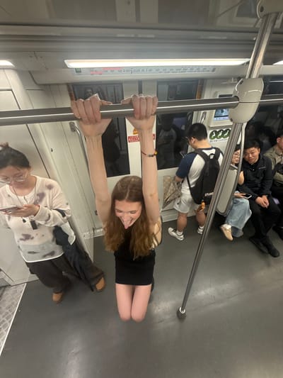 A young Caucasian woman with long, wavy brown hair is hanging from a horizontal metal bar inside a subway car in Changning District, China. She is wearing a sleeveless black dress and has her tongue sticking out. Her arms are extended upwards, gripping the bar. To her left, an East Asian woman wearing glasses and a tie-dye-patterned shirt is looking down at her phone. Other passengers are seated and standing in the subway car, with some visible in the background. The subway car is illuminated by fluorescent lights and displays overhead information screens. The floor is dark gray and appears to be made of a durable material.