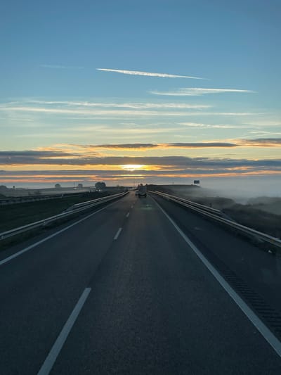 An open highway stretches into a sunrise horizon. The sky is filled with long, vertical streaks of clouds reflecting the sun's golden glow. A single contrail cuts diagonally across the clear blue portion of the sky. The asphalt road is gray, marked with white lines, and bordered by metal guardrails. A few vehicles can be seen further down the highway.