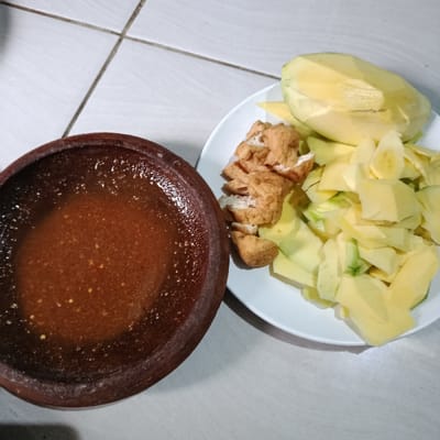 A white plate holds peeled and sliced mangoes, some cut into chunks and others into thin pieces, alongside fried tofu cubes. Next to it, a dark brown clay bowl is filled with a reddish-brown liquid, possibly a spicy sauce, speckled with visible chili seeds. The arrangement sits on a surface of white tiles, creating a casual, potentially homemade food display.