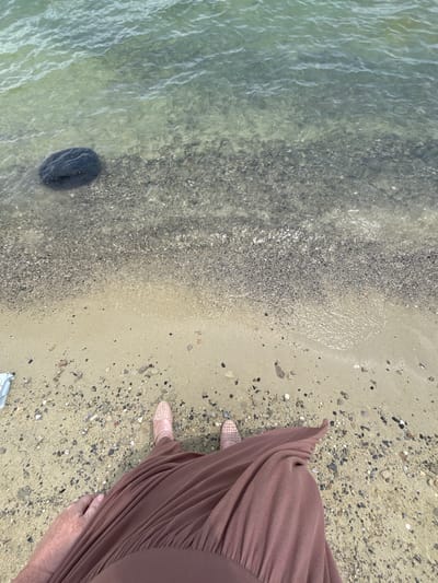 The image depicts a first-person view from a person standing on a sandy beach near the water's edge. The water is clear and shallow, with small pebbles and sand visible on the seabed. A large dark rock is submerged in the water to the left of the frame. The person is wearing a long, brown, pleated skirt and light pink woven shoes. The beach is covered in sand and a mixture of small pebbles and shells. The overall scene suggests a serene coastal environment, possibly in the city of Tsamakaberd, Armenia, known for its coastline along Lake Sevan.