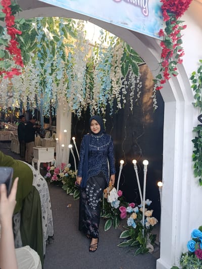 The image captures a young adult woman standing beneath a decorative archway at an indoor/outdoor event in Kedung Dowo, Indonesia. The woman is positioned slightly right of center, facing forward with a smile towards the camera. She wears a dark blue traditional long-sleeved top (kebaya-style) adorned with intricate white beadwork around the neckline and cuffs, and a matching dark blue hijab. A blue shawl is draped over her left shoulder. Her long skirt is dark blue with white and silver floral and bird patterns, consistent with batik. She holds a small white and gold structured handbag in her right hand and wears black open-toed sandals.
The archway is white and multi-layered, lavishly decorated with cascades of artificial white and light blue wisteria-like flowers, interspersed with variegated green and white leaves hanging from the top. Red floral garlands are draped over the right side of the arch, with additional green leafy vines trailing down the right pillar.
At the base of the arch, several white poles topped with spherical lights extend upwards, surrounded by arrangements of artificial pink, blue, and yellow roses with green foliage. The ground is a dark gray textured surface. Behind the woman, a dark backdrop is illuminated by warm string lights. To her left, beyond the floral arrangements, an event space is visible with tables covered in white tablecloths and chairs. Two men are present in the background; one in a dark green uniform stands near a table, and another in a black shirt holds a camera, facing away from the viewer. A small white cabinet is also visible near the tables. A banner, partially visible at the top of the arch, displays light blue and purple text, possibly reading "Dung" as part of a larger word or name.