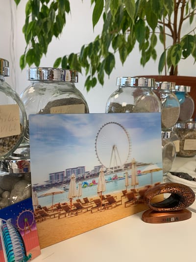 The image depicts a close-up, eye-level view of a white surface, likely a table, upon which several objects are arranged. In the foreground, a small, bronze-colored souvenir sits on a base that reads "DUBAI." The souvenir is an oval shape with intricate etched patterns and Arabic calligraphy. To the left of the souvenir, a rectangular postcard is propped up, displaying a photograph of a beach scene with lounge chairs, umbrellas, and a large Ferris wheel in the background, also featuring the text "DUBAI."
Behind the postcard, several glass jars are visible, arranged in a row. These jars have silver lids and contain various granular substances, possibly sand or small stones. One jar on the far left has a handwritten label with illegible text. Another jar in the center has a label that reads "kemer."
In the background, a large green leafy plant is prominent, its branches extending across the top of the frame. The wall behind the plant is white. The overall impression is that of a collection of travel souvenirs and natural elements displayed on a flat surface in an indoor setting. The location is identified as the city of Kommunar, Russia.