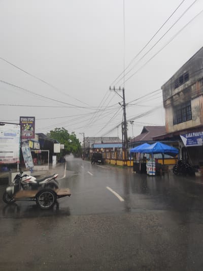 This photograph captures a quiet street scene in Keudematangpanyang, Indonesia, under an overcast sky. The wet asphalt road, reflecting the dull light, suggests recent rainfall or ongoing light drizzle. The overall atmosphere is subdued and calm.
On the left side of the street, a white and red motorcycle with a wooden sidecar is parked near the foreground. Behind it, several commercial signs are visible, including a prominent "SYARA STORE" sign, which also mentions "TANAH PASER" and "MULAI DARI 10 RIBUAN" (starting from 10,000). Other unreadable posters and signs are affixed to structures along this side. Numerous tangled overhead utility lines crisscross the sky, supported by a tall utility pole. Further down, buildings with brown roofs and lush green trees line the road.
The right side of the street features a multi-story building, part of which is occupied by a business named "ZARA PARFUM." Below this name, services like "PULSA," "VOUCHER," "TOKEN LISTRIK," and "GoPay" are advertised. In front of this building and extending further down, two blue canopies or tents provide shelter, possibly for vendors or customers. Other motorcycles are parked closer to these tents. The road itself has a faded white line marking its center.
No people are visible, contributing to the tranquil appearance of the street. The heavy, grey sky dominates the upper portion of the image, indicating a cloudy or rainy day.