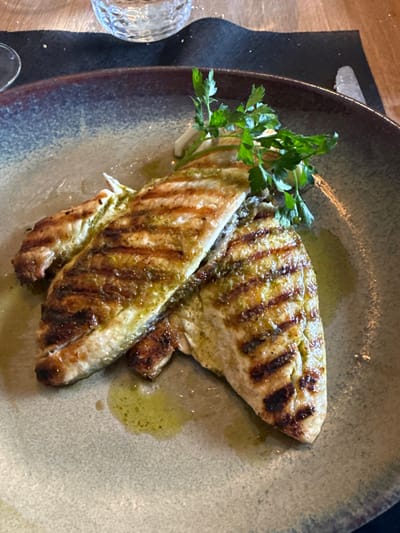 The image displays a close-up overhead view of two grilled fish fillets served on a round, speckled ceramic plate with a raised rim. The fillets exhibit prominent dark brown char marks from grilling and are coated with a bright green herb-based oil or sauce. A small cluster of fresh green parsley sprigs garnishes the fish towards the top center of the plate. Olive oil is visible pooling around the fillets. In the background, a portion of a clear, textured glass is visible on a dark gray or black fabric placemat, which rests on a light brown wooden table. A silver knife handle is partially visible on the right edge of the placemat. The scene depicts a meal served in a restaurant setting in Mora, Portugal. No people are visible.
