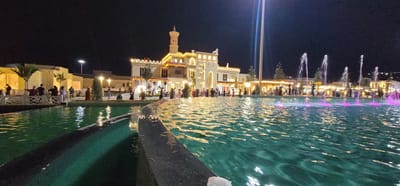 In the city of Satkak, Uzbekistan, at night, a lively scene unfolds around a large, illuminated fountain. The fountain's water is a vibrant turquoise, reflecting the surrounding lights, and several jets of water shoot upwards, some with a pinkish glow. Behind the fountain, a crowd of people is gathered, observing the spectacle.
The background features a distinctly architectural style, with buildings that appear to be made of light-colored stone, possibly stucco. One prominent building is a multi-story structure with a tower that has a distinct, onion-shaped dome, reminiscent of Islamic architecture. This building is brightly lit with yellow and white lights, highlighting its intricate details, including arched windows and decorative elements along the roofline. Other buildings in the background are lower in height and also adorned with decorative lighting, creating a festive atmosphere.
The ground in front of the buildings and around the fountain appears to be paved. Small trees with sparse foliage are scattered throughout the scene, and a tall, slender pole, possibly a light pole, stands prominently to the right of the main building.
The people in the image are dressed in casual attire and appear to be enjoying the evening. Some are standing, some are sitting, and some are walking. The overall impression is one of a public gathering space, perhaps a park or a plaza, where locals and possibly tourists come to relax and enjoy the illuminated water features and architecture. The darkness of the night sky provides a stark contrast to the bright lights of the fountain and the buildings, making the scene visually striking.