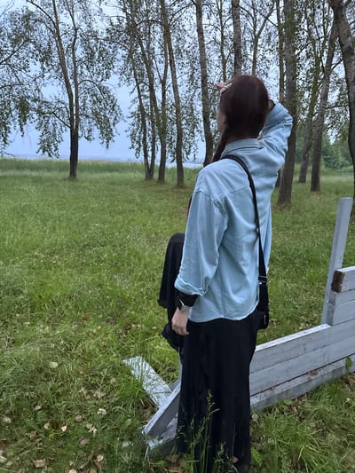A young woman stands in a grassy field, facing away from the camera, looking towards a body of water and a line of birch trees under a cloudy sky. She has dark hair in a braid and is wearing a light blue collared shirt, a black skirt, and a black crossbody bag. She is holding her hand up to her forehead, shielding her eyes from the sun. A white wooden fence is visible in the foreground to the right. The overall scene suggests an overcast day in a natural, possibly park-like setting, consistent with the outskirts of Nevyansk, Russia, a city known for its historical architecture and proximity to natural landscapes.