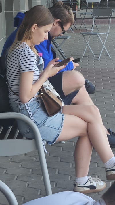 A young woman with blonde hair and a striped shirt sits on a bench next to a man with brown hair and glasses. She is looking at her phone, which she holds in her hands. The man is also looking at his phone. They are both sitting outdoors on a paved area with some chairs in the background. The image appears to have been taken in the city of Kysak, Slovakia.