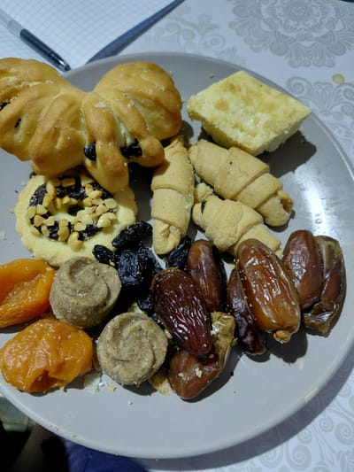 A plate of assorted sweets and dried fruits is displayed on a table with a patterned tablecloth. The plate contains two butterfly-shaped pastries with raisins, a small tart topped with chocolate and nuts, several crescent-shaped cookies, dried apricots, round, swirled sweets, raisins, and dates. A notepad and two pens are visible in the upper left corner. The scene appears to be indoors.