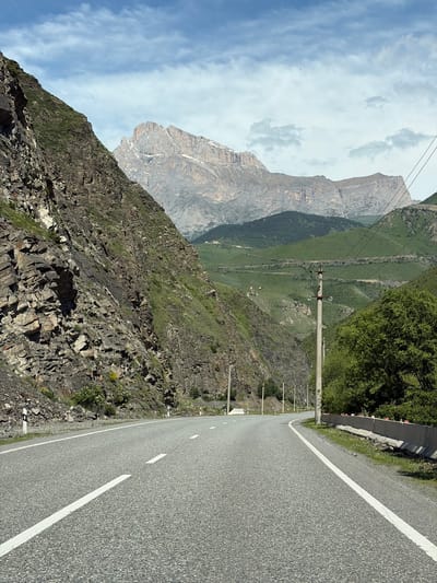 The image captures a winding asphalt road through a mountainous valley. On the left, a steep, rocky mountainside covered in sparse vegetation dominates the foreground. The road curves gently to the right, bordered by a concrete barrier with red reflectors on the right side. In the distance, rolling green hills ascend to a rugged, snow-capped mountain peak under a partly cloudy blue sky. Power lines are visible, supported by concrete poles, traversing the landscape. The scene appears to be a scenic route in Nizhnyaya Unal, Russia, characterized by its dramatic natural terrain and infrastructure for travel.