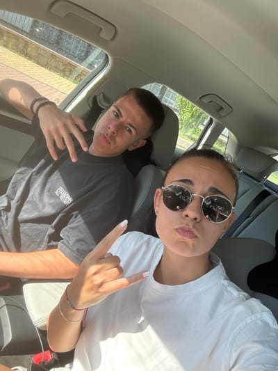 In this photo, a young man and woman are seated in the interior of a car, likely on a sunny day, judging by the bright lighting and shadows. The man is in the foreground, looking towards the camera with a neutral expression. He is wearing a dark t-shirt with white text that reads "PEGADOR ALL WE GOT". He is making a peace sign with his left hand and has a bracelet on his right wrist. The woman is positioned behind and to the right of the man, also looking towards the camera. She is wearing a white t-shirt and large, round sunglasses. Her lips are pursed, giving her a playful or pouting expression. She is making a rock-on hand gesture with her right hand, which is adorned with two bracelets. The background shows the interior of a car, including seats, a handle on the ceiling, and a window that offers a glimpse of green foliage and what appears to be a stone wall or structure outside, suggesting an outdoor setting, possibly Vrachesh, Bulgaria. The overall mood of the image appears casual and friendly, with the two individuals capturing a moment together in the car.