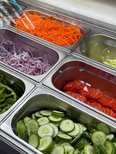 The scene depicts a close-up, overhead view of a food preparation or serving station, likely a salad bar, in ตำบล ไม้ขาว, Thailand. Six stainless steel food pans, or Gastronorm containers, are arranged within a larger metal frame.
In the top left container, a quantity of finely shredded orange carrots is present. Partially covering this container is a clear, reflective lid. Adjacent to this, in the top right, another stainless steel container holds a translucent, pale yellow liquid containing small green fragments, possibly a dressing or brine.
Below the carrots, to the left, a pan contains thinly sliced red onions. Below the red onions, to the left, another pan holds chopped green bell peppers.
Below the liquid container, to the right, a pan is filled with neatly arranged, thinly sliced red tomatoes. The interior of this pan exhibits a reddish hue. Below the tomatoes, at the bottom right of the frame, a pan contains a collection of freshly cut green cucumbers, comprising both round slices and longer, quartered pieces.
The metal surfaces of the containers and framework appear clean, with minor reflections from overhead lighting visible on the clear lids and metal. No individuals are visible in the frame, and no specific actions or interactions are occurring. The scene presents a static display of prepared salad ingredients.
