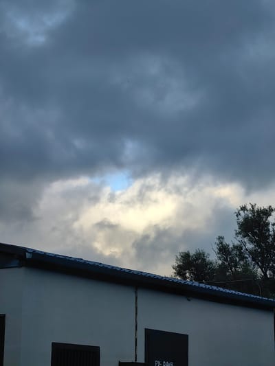 The image captures a cloudy sky over a building with a dark, corrugated roof. The building is light-colored with dark rectangular window or door openings visible on its facade. Trees are partially visible on the right side of the frame, silhouetted against the dramatic sky. The overall atmosphere suggests overcast weather, possibly before or after rain, in an urban or semi-urban setting. The location is identified as Фаніпальскі сельскі Савет, Belarus.