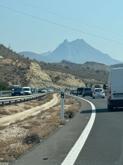 A highway scene in Cala D'or, Spain, features a moderate flow of traffic on a sunny day under a clear blue sky. The road curves to the right, bordered by a guardrail and a dry, scrub-covered embankment on the left. On the right, a white van is prominently in the foreground. The distant landscape is characterized by arid, layered hills leading up to a prominent, jagged mountain peak under a hazy sky. Several cars and vans are visible in both lanes of traffic.