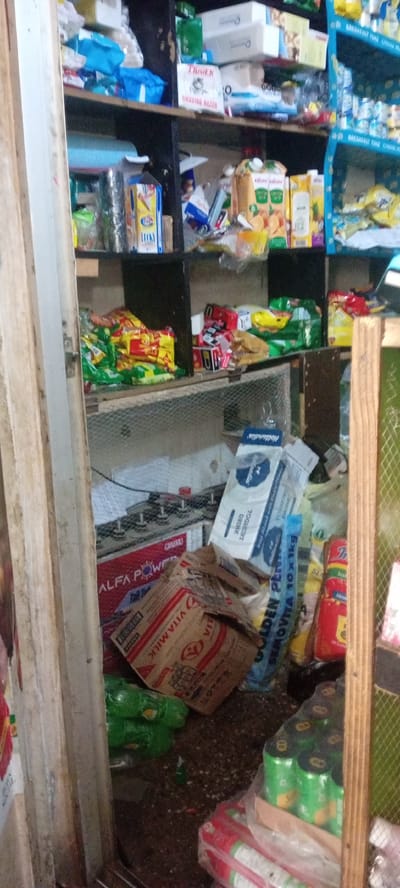 The image depicts a dimly lit, cluttered storage area or small shop in Kwachiri, Nigeria. Shelves are packed with various items, including packaged food products, boxes, and what appears to be electronic equipment. On the lower shelves, there are bags of snacks, cartons of juice, and a large battery or power supply unit. There are also items on the floor, including green plastic bags and several stacks of canned drinks. The overall impression is one of disarray and limited space, typical of a small retail or storage environment.