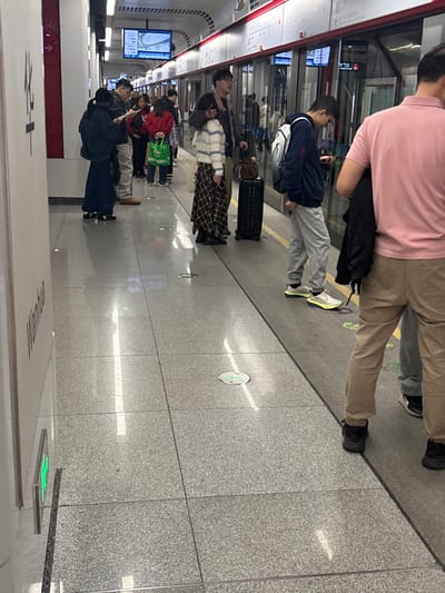 People stand in line on a subway platform waiting for a train. The floor is tiled and has round markers with arrows on them. The train doors are closed. Some of the people have luggage. The angle of the photo is sideways, with the platform taking up most of the space. The subway car is white and red. There are digital displays with schedules inside the train.