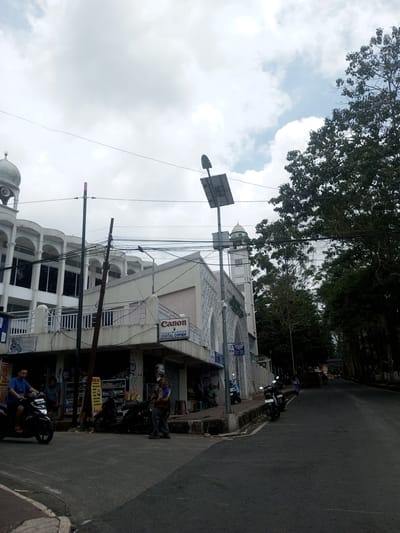 The image captures a street scene in Ban Phouviengnoy, Laos. The intersection is dominated by a multi-story building, primarily white, with architectural elements including arches and a dome. A "Canon" sign is visible on the building's side, suggesting a business. Power lines crisscross overhead. On the street, a man rides a scooter, while another stands near parked motorcycles. The road stretches into the distance, flanked by trees. The sky is overcast.