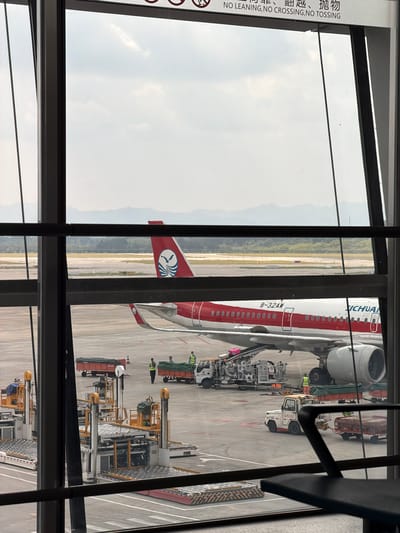 The image shows an airport tarmac with a Sichuan Airlines airplane parked at a gate. The airplane is red and white with the Sichuan Airlines logo on its tail. Various ground support equipment, including baggage carts and a tug, are visible around the aircraft. Several airport workers are present, some attending to the equipment and others near the plane. The scene is viewed through an airport terminal window, with the sky and distant mountains visible in the background. The text on a sign above the window indicates "NO LEANING, NO CROSSING, NO TOSSING." The location is Jianyang City, China.
