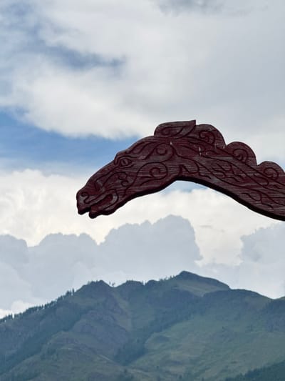 An outdoor, eye-level shot captures a detailed, carved wooden dragon head, painted a deep red, extending from the right side of the frame, its snout pointing left. The dragon's head features intricate swirling patterns carved into its surface, and its eye is a circular indentation. The background is a layered landscape of sky and mountains. The upper portion of the sky is a pale blue with wispy white clouds. Below the sky, a range of rugged, green mountains stretches across the horizon, dotted with dark green trees on their slopes. The overall lighting suggests it is daytime, with diffused light illuminating the scene. The image is sharp and clear, focusing on the dragon head's texture and the mountain's form. The location is Anchyl-Chon, Russia.