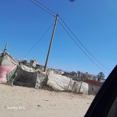 The image captures a street scene in the city of Deir el-Balah, Palestinian Territories. In the foreground, a sandy ground is littered with debris. Behind it, a makeshift fence constructed from tarps, netting, and barbed wire encloses an area. A wooden utility pole with electrical wires and a single lightbulb stands prominently on the left side of the frame. In the background, several buildings are visible, including residential structures and what appears to be an unfinished building with exposed rebar. A palm tree is also present. The sky is a clear, bright blue. The overall impression is one of a developing or possibly impoverished urban environment.