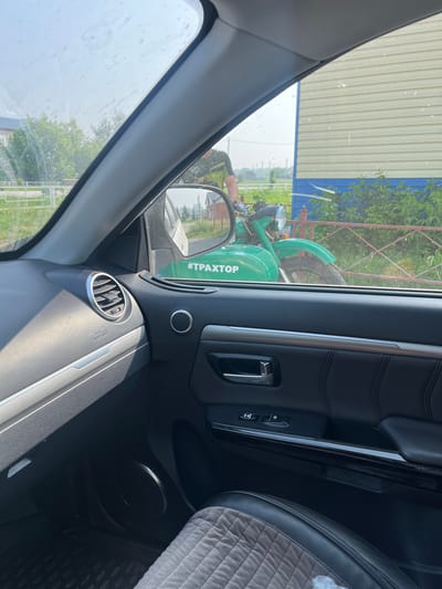 The image shows a view from inside a car in the city of Gorskin, Russia. Through the passenger side window, a green motorcycle is visible parked in front of a building with yellow siding and a blue stripe. The motorcycle has the hashtag "#ТРАХТОР" written on its side. A person wearing a green and black shirt is partially visible standing behind the motorcycle, appearing to be the rider. The environment outside the car suggests a rural or suburban setting with green foliage and a fence. Inside the car, the dashboard, door panel, and a textured seat cover are visible, indicating the perspective is from the driver's or passenger's seat. The sky is bright, suggesting daylight.
