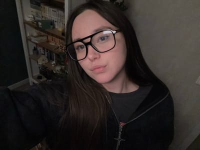 An indoor scene in Prikubanskiy, Russia, depicts a female subject captured in a close-up, chest-up selfie. The subject is a woman with long, dark brown hair parted on the left side, extending over her shoulders. She wears black-framed eyeglasses, a dark gray t-shirt, and an unzipped black hooded jacket with a silver zipper and a silver four-pointed star pendant visible on the zipper pull. Her expression is neutral, with slightly pursed lips.
The background to the left of the subject reveals a white-framed multi-shelf unit, likely a bookshelf, containing various items including books, a green box, and assorted decorative objects. A dark green wall section is visible further to the left. The background to the right of the subject consists of a plain light gray wall with a white electrical outlet visible in the lower right corner. The lighting suggests an indoor, ambient setting.