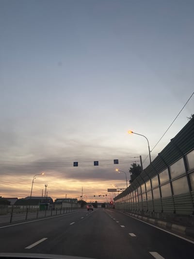 This image depicts a road in Mstikhino, Russia, at dusk or dawn. The sky is a pale blue with wispy clouds, transitioning to warmer hues of orange and yellow near the horizon. A road stretches into the distance, marked with white dashed lines. On the right side of the road is a tall, green sound barrier constructed of panels. Streetlights with orange lamps are visible along the roadside, with some illuminated. Traffic signs, including directional arrows, are suspended over the road on wires. On the left side, there are buildings and houses with green roofs, and a cellular tower is visible in the distance. A single vehicle, a silver SUV, is driving away from the camera on the right lane of the road. The overall atmosphere is serene, with the dimming light suggesting either the end of the day or the beginning of a new one.