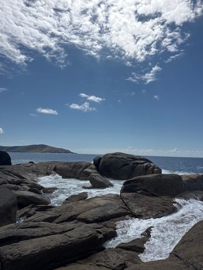 The image shows a rocky coastline in Muxía, Spain, at latitude: 43.113, longitude: -9.221. The scene is composed of large, gray rocks and white foamy waves crashing on the shore. The sky is clear blue with white cumulus clouds. There is a distant green, hilly landmass visible on the horizon. The rocks are scattered across the shore, with some forming a barrier to the water. The waves are breaking on the rocks, creating a white, foamy spray.  There are no people or animals present in the image.