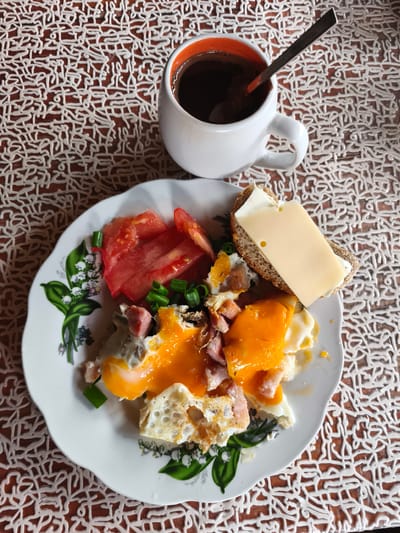 The image shows a close-up, top-down view of a breakfast spread on a wooden table covered with a white, textured placemat. The meal consists of a white plate with a floral pattern, holding scrambled eggs with melted cheese, chopped ham, sliced tomatoes, and chopped green onions. To the right of the plate is a slice of bread topped with a slice of cheese. A white mug with an orange interior, filled with dark coffee, is positioned above the plate, with a spoon resting inside. The background is a dark, wooden surface with a distinct white, lace-like placemat. The scene appears to be set indoors. The image does not contain any elements that would allow for the determination of its location in Dolzhikovo, Russia.