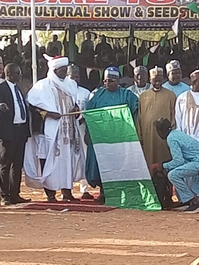 An outdoor shot features a group of men at an "Agricultural Show & Seeds" event. Most wear traditional clothing; one is in a suit. A man in white robes and a red fez holds a Nigerian flag. Another man in green stands beside him. A banner is visible in the background, with more people behind it. A person in blue kneels near the flag's edge. The setting appears to be a sandy or dirt surface.