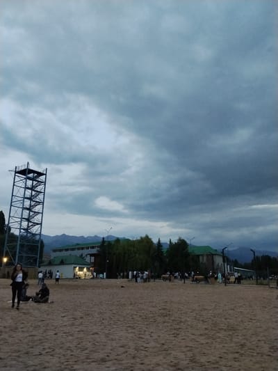 In Bosteri, Kyrgyzstan, a group of people are gathered on a sandy area near a body of water. In the foreground, a woman in black pants and a white top walks across the sand, while another person sits on the ground nearby. Further back, several individuals are seen standing and walking. To the left of the frame is a tall, metallic observation tower with stairs. Behind the people and tower, a long building with a green roof is visible, with more people milling about in front of it. Trees line the area behind the building, and mountains are visible in the distance under a cloudy sky. The scene appears to be a public gathering space, possibly a park or a recreational area by a lake.