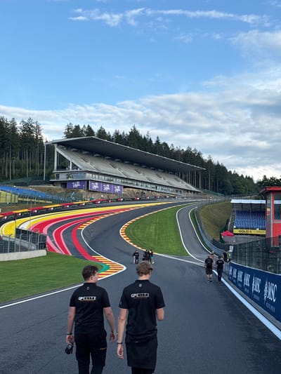 An outdoor, wide-angle image depicts the Circuit de Spa-Francorchamps in Stavelot, Belgium, under a partly cloudy sky. The asphalt racetrack, identified by its distinctive red, yellow, and black (with added pink) painted run-off area on the left, curves from the foreground towards a steep uphill section, characteristic of the Eau Rouge/Raidillon complex.
In the foreground, two individuals, viewed from behind, walk on the left side of the track. Both wear black short-sleeved shirts with "GPELITE DRIVE TO PERFECTION" visible in white text on their backs. The person on the left holds black gloves in their right hand.
Further along the track, several other individuals, similarly dressed in dark clothing, are walking. To the left of the track, a large, multi-tiered concrete grandstand with a gray roof is positioned on a sloped embankment, partially obscured by green fencing. Purple banners bearing the "Virgin" logo and other markings are visible beneath its seating area. Directly across from this grandstand, on the right side of the track, a blue advertising barrier featuring "MSC CRUISES" branding runs parallel to the track. Behind this barrier, a smaller blue grandstand, labeled "GOLD 5," is visible with red support structures.
Dense green pine forests line both sides of the track and rise on the hills in the background. The sky above is a mix of clear blue and scattered white cumulus clouds.