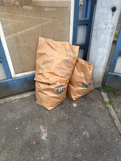 The image shows two brown paper yard waste bags labeled "COLLECTES DES DECHETS" with a green and blue logo, possibly related to the city of Orsay, France, where the bags are likely used for municipal yard waste collection. The bags are placed near a blue and silver metal fence or gate on a concrete surface. Text on the bags includes instructions such as "Ne fermez pas vos sacs" and lists acceptable and unacceptable items for composting, such as grass clippings and flower waste. The location appears to be an outdoor area, potentially a curbside or a designated collection point.