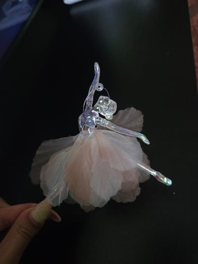 This image depicts a translucent, iridescent ballerina ornament held by a human hand against a dark, flat surface, likely a table or desk, in Staliyska mahala, Bulgaria. The ornament, appearing to be crafted from glass or acrylic, features a ballerina in an arabesque pose with one arm raised and the other extended, and one leg extended backward. A layered tutu, composed of light pink or white tulle, is affixed to the ballerina's midsection. A small metallic hanging loop is visible near the figure's raised arm. A human hand, positioned in the lower left quadrant of the frame, with almond-shaped fingernails painted with glittery light green polish, is grasping the lower section of the ballerina's extended leg. The environment is dark and appears to be indoors.