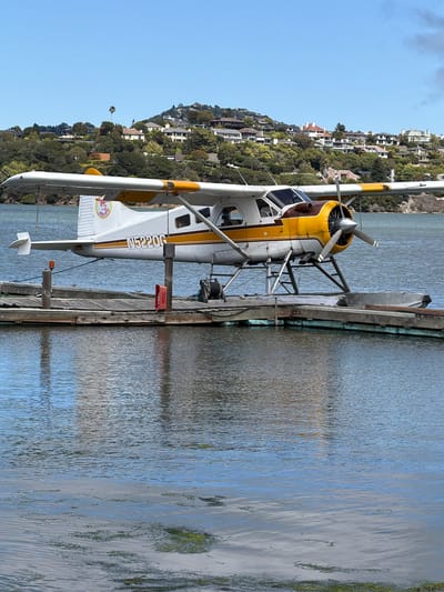 A white and yellow floatplane, identified by the registration number N5220G, is docked at a wooden pier on a body of water. The airplane has a single engine and a propeller. The background features a lush green hillside dotted with houses. The sky is clear and blue. The scene is likely located in Marin City, United States of America, given the distinctive architecture and landscaping often found in the region. There are no people visible in the image. The pier is weathered and shows signs of use, with a red fire extinguisher and a coiled rope visible on its surface.