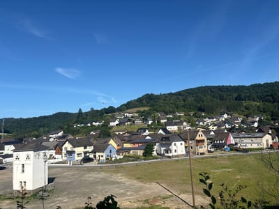 The image shows a view of the town of Schuld, Germany, with houses built on a hillside. The sky is clear and blue, with a few wispy clouds. The foreground is a dirt and gravel area with some green grass and small plants. In the middle ground, there is a street lined with various houses, some with solar panels on their roofs. A white, rectangular building with windows and what appears to be electrical equipment is prominent in the left foreground. Behind the houses, the hillside is covered in lush green trees. The architecture of the houses suggests a European village setting. There are no people visible in the image.
