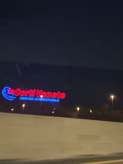 The image shows a neon sign that reads "le Corti Venete CENTRO COMMERCIALE" in red and blue lettering. The sign is illuminated against a dark night sky. In the background, there are several streetlights and a low wall. The location is identified as San Martino Buon Albergo, Italy. The sign appears to be for a shopping center. There are no people visible in the image.