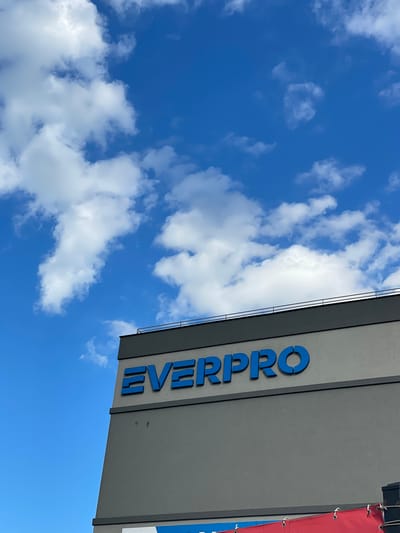 The image shows a partly cloudy blue sky next to the corner of a building. The side of the building is painted grey and has the word "EVERPRO" vertically displayed in blue letters. The building appears to be in Afumați, Romania. A small portion of a red and blue banner is visible on the right side of the building.