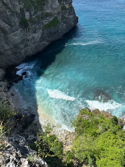 A high-angle shot depicts a coastal scene in Pejukutan, Indonesia. Dominating the left and right sides are rugged cliffs, with the left one appearing darker and more textured, showing various geological layers. The right cliff is covered with dense green vegetation. The water between the cliffs showcases varying shades of blue, from a lighter turquoise to a deep blue, influenced by sunlight and depth. White waves crash against the shore and the base of the cliffs, indicating active water movement. A small sandy beach is visible at the bottom center. The perspective suggests the shot was taken from a high vantage point, likely overlooking the coastline. No people are visible in the scene.