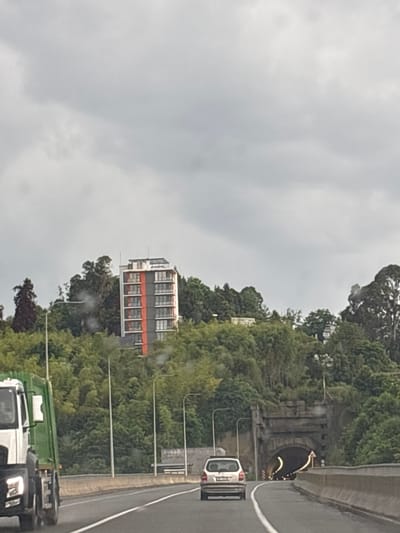 The image depicts a cloudy day scene on a highway in Makhinjauri, Georgia, with a distinct focus on a multi-story building and a tunnel entrance. In the foreground, a white and green truck is partially visible on the left, moving along the road. Further ahead on the right side of the highway, a silver hatchback car is visible. Dominating the background on a hillside covered in dense green foliage is a modern, tall building with white and orange vertical accents, featuring multiple balconies and windows. To the right of the building, an old stone tunnel entrance is situated within the greenery, with lights visible inside and traffic signage nearby. The sky is overcast with gray clouds.