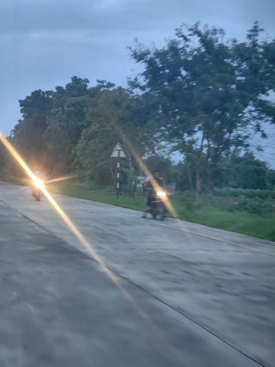 The image depicts a two-lane road, likely in Padli, India, with a motorcyclist in the foreground on the left, moving away from the camera. The motorcyclist's headlight is intensely bright, creating a strong lens flare that dominates the left side of the image and extends diagonally across the road. The road surface appears to be asphalt or concrete and is slightly wet.
To the right of the road, there is a strip of green grass followed by a dense line of trees and foliage. The sky is overcast and a pale blue-grey, indicating either dawn, dusk, or a cloudy day. A triangular road sign with a black border and white interior, with a black symbol, is visible on a pole to the right of the road, near the trees.
A second motorcyclist is visible further down the road on the right, also moving away from the camera. This motorcyclist's headlight is also illuminated, though less intensely than the first. The overall impression is of a rural or semi-rural setting on a road with active traffic, captured in low light conditions.